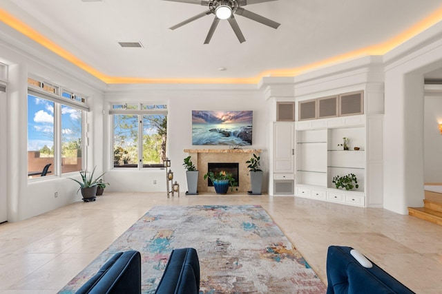 living room with a raised ceiling, a high end fireplace, light tile patterned floors, built in shelves, and ceiling fan