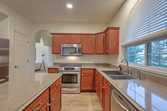 kitchen featuring arched walkways, light wood finished floors, appliances with stainless steel finishes, a sink, and light stone countertops