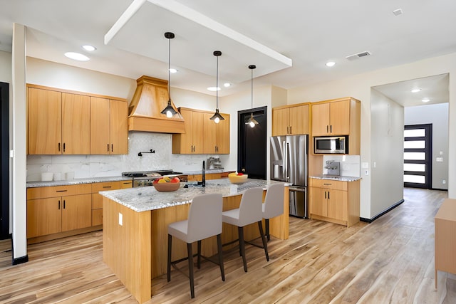 kitchen featuring a center island with sink, premium range hood, stainless steel appliances, light stone counters, and light wood-type flooring