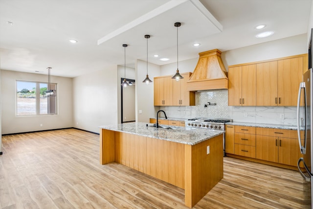 kitchen featuring custom range hood, light wood-type flooring, light stone counters, an island with sink, and pendant lighting