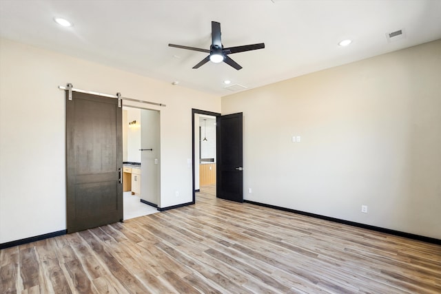 unfurnished bedroom featuring ensuite bathroom, ceiling fan, a barn door, and light hardwood / wood-style floors