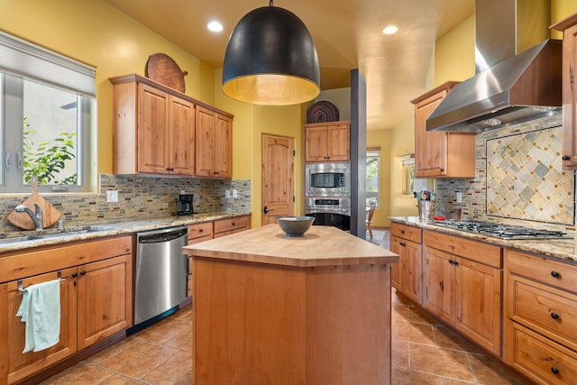 kitchen featuring wood counters, backsplash, appliances with stainless steel finishes, a center island, and wall chimney range hood