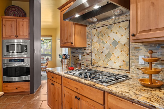 kitchen featuring backsplash, appliances with stainless steel finishes, light stone counters, wall chimney exhaust hood, and light tile patterned flooring