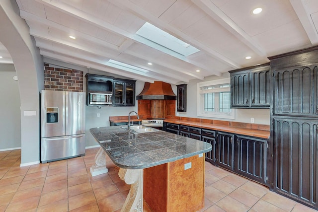 kitchen featuring vaulted ceiling with skylight, a kitchen island with sink, sink, appliances with stainless steel finishes, and premium range hood