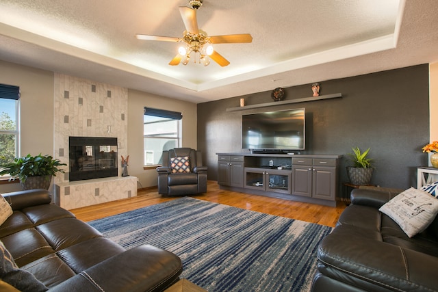 living room with a textured ceiling, a fireplace, light hardwood / wood-style flooring, a raised ceiling, and ceiling fan