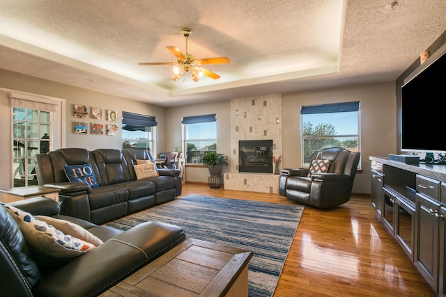 living room with light wood-type flooring, a tray ceiling, a textured ceiling, a tiled fireplace, and ceiling fan