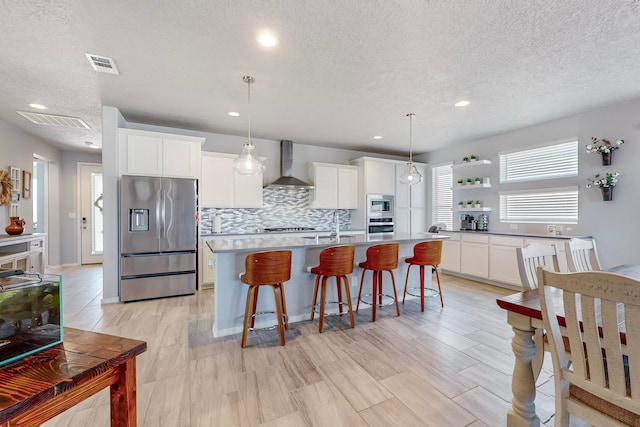 kitchen with stainless steel appliances, wall chimney exhaust hood, decorative light fixtures, white cabinets, and a kitchen island with sink