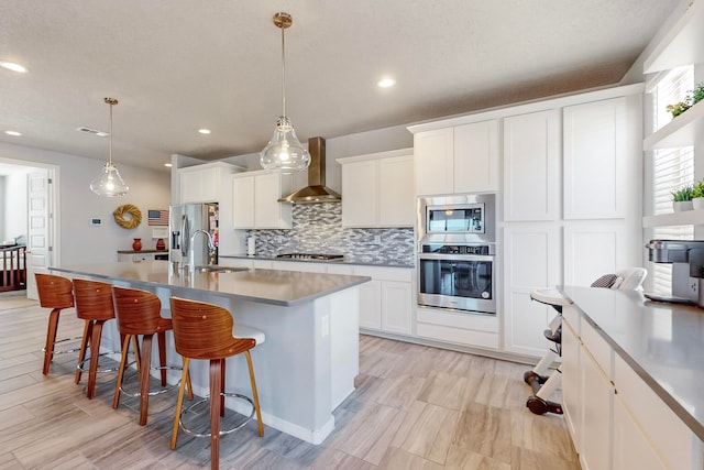 kitchen featuring appliances with stainless steel finishes, a kitchen island with sink, decorative light fixtures, and wall chimney range hood