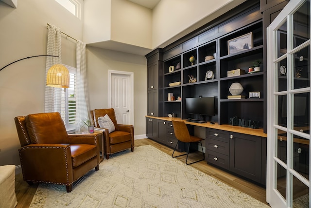 sitting room with built in desk, light wood-type flooring, a towering ceiling, and baseboards