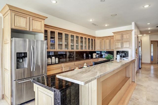 kitchen with light brown cabinetry, stainless steel fridge, tasteful backsplash, a kitchen island with sink, and dark stone countertops