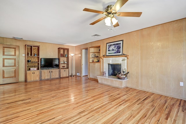 unfurnished living room featuring ceiling fan, built in features, wooden walls, a tile fireplace, and light hardwood / wood-style floors