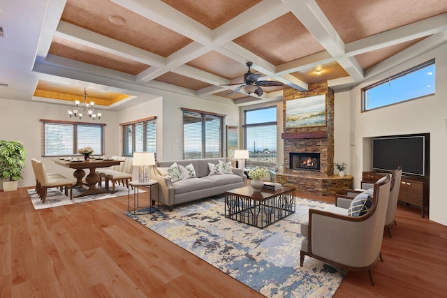 living area with coffered ceiling, a high ceiling, a stone fireplace, light wood-style floors, and ceiling fan with notable chandelier