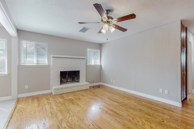 unfurnished living room with a brick fireplace, wooden walls, ceiling fan, light hardwood / wood-style flooring, and a textured ceiling