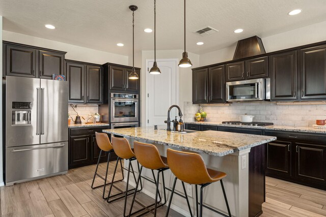 kitchen featuring an island with sink, hanging light fixtures, sink, light hardwood / wood-style flooring, and stainless steel appliances