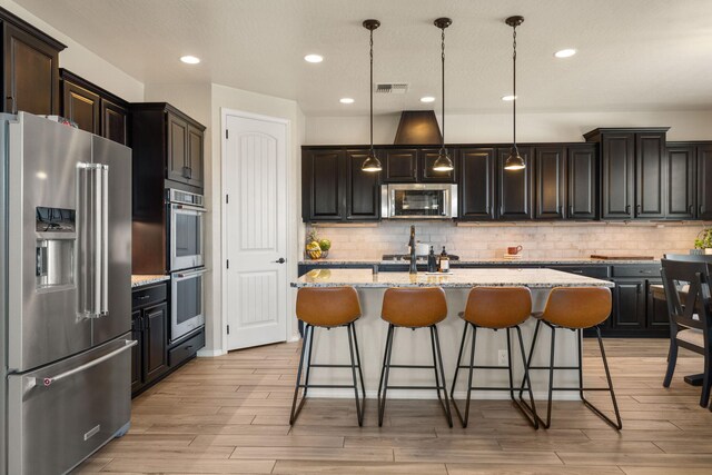 kitchen featuring light stone counters, a kitchen island with sink, stainless steel appliances, hanging light fixtures, and light hardwood / wood-style flooring