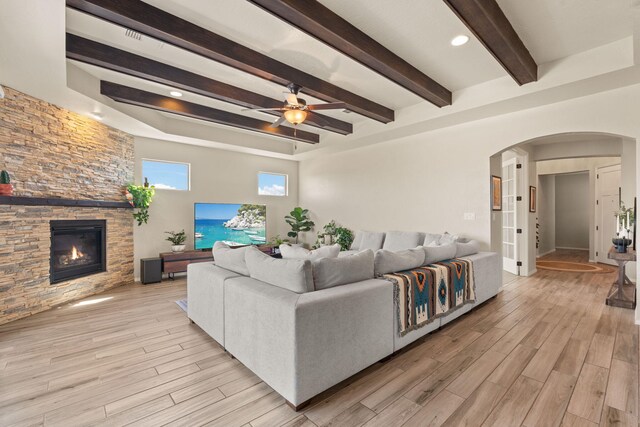 living room featuring light wood-type flooring, beamed ceiling, ceiling fan, and a stone fireplace