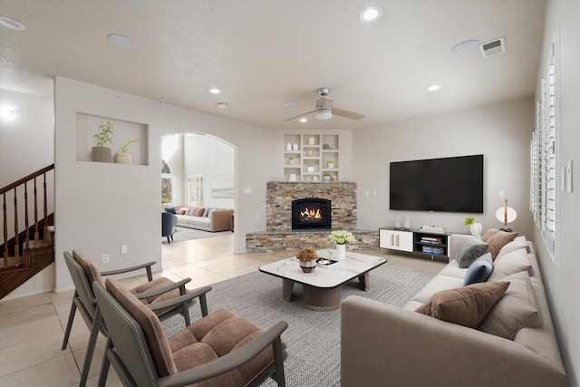 tiled living room with built in shelves, ceiling fan, and a stone fireplace