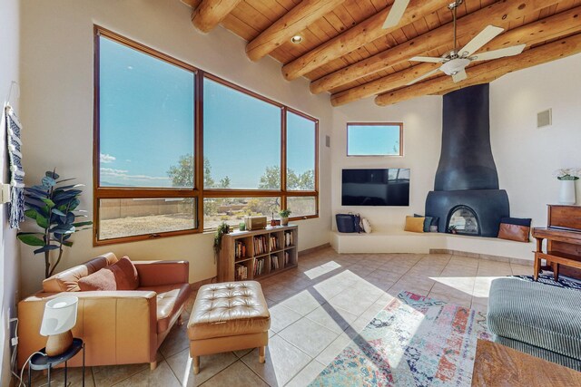 living room with ceiling fan, wooden ceiling, beamed ceiling, a wood stove, and light tile patterned flooring