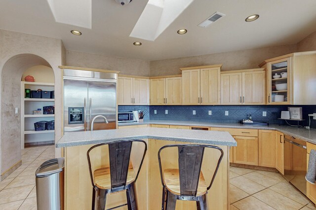 kitchen with a skylight, light brown cabinets, light tile patterned floors, built in refrigerator, and an island with sink