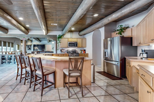 kitchen featuring appliances with stainless steel finishes, light brown cabinetry, a kitchen bar, beam ceiling, and light tile patterned floors