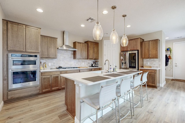 kitchen with wall chimney exhaust hood, hanging light fixtures, a kitchen breakfast bar, a center island with sink, and appliances with stainless steel finishes