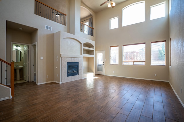 unfurnished living room with a towering ceiling, built in features, a tile fireplace, ceiling fan, and dark wood-type flooring