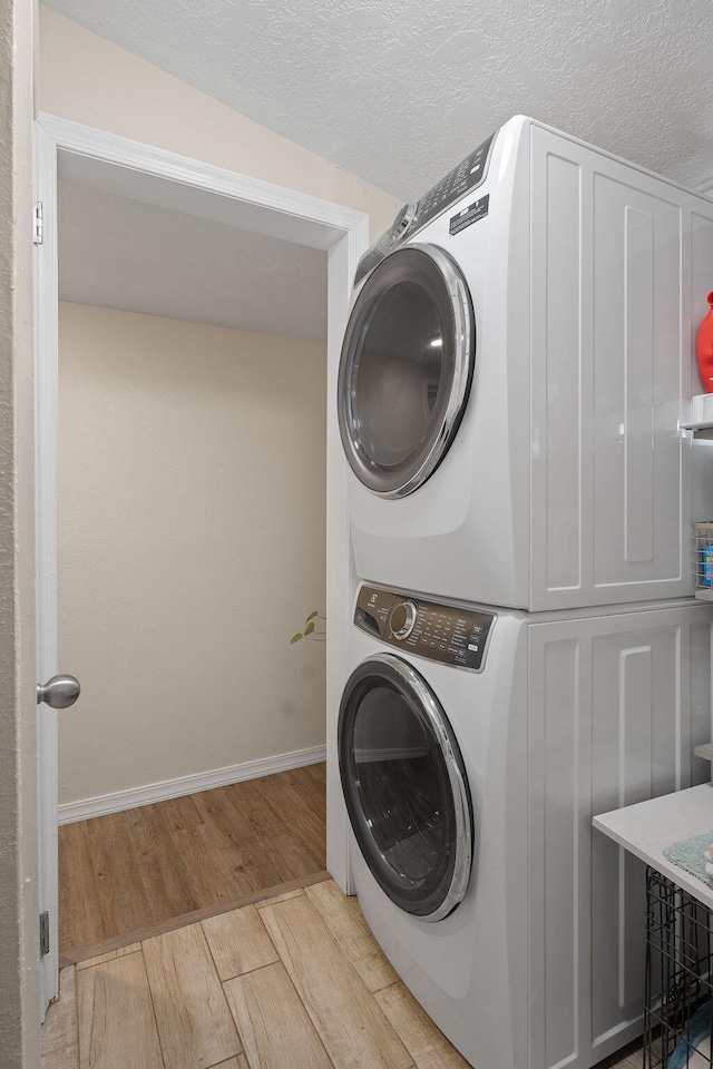 clothes washing area featuring light wood-style flooring, a textured ceiling, stacked washing maching and dryer, laundry area, and baseboards