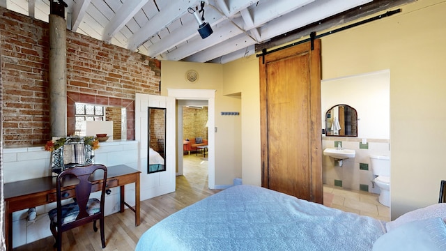 bedroom with a barn door, connected bathroom, brick wall, light wood-style flooring, and beam ceiling