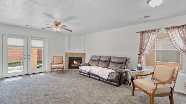 carpeted living room featuring ceiling fan, a tiled fireplace, a textured ceiling, and french doors