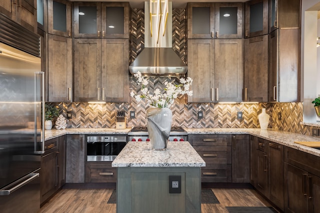 kitchen featuring tasteful backsplash, stainless steel appliances, wall chimney exhaust hood, dark wood-type flooring, and light stone counters