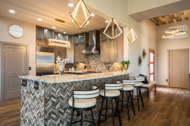 kitchen with wall chimney exhaust hood, built in fridge, light stone countertops, dark hardwood / wood-style flooring, and tasteful backsplash