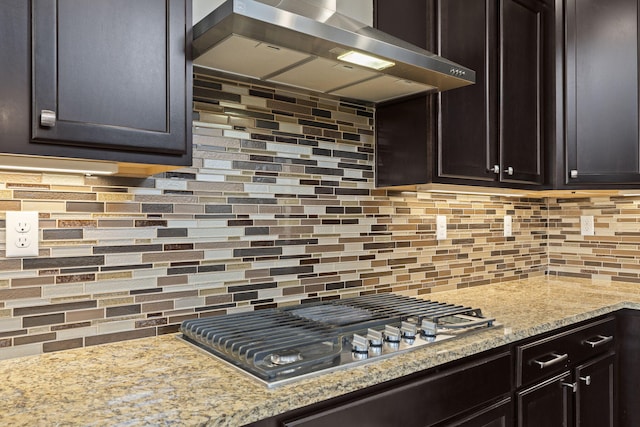 kitchen with stainless steel gas cooktop, wall chimney exhaust hood, dark brown cabinetry, light stone counters, and tasteful backsplash