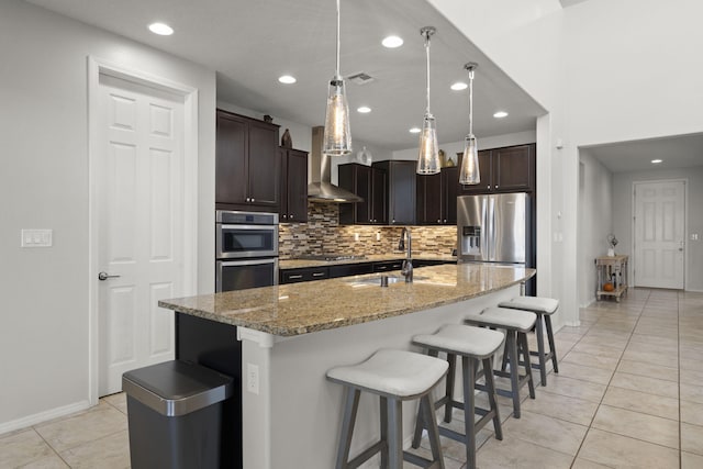 kitchen featuring an island with sink, stainless steel appliances, sink, light stone countertops, and dark brown cabinetry