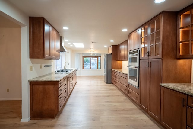 kitchen with a skylight, light stone counters, wall chimney exhaust hood, stainless steel appliances, and light hardwood / wood-style flooring