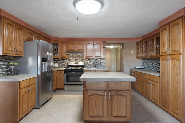 kitchen featuring brown cabinetry, stainless steel appliances, light countertops, glass insert cabinets, and under cabinet range hood