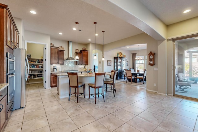 kitchen featuring wall chimney range hood, backsplash, decorative light fixtures, stainless steel refrigerator, and a kitchen island with sink