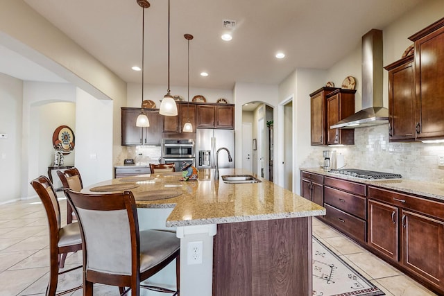 kitchen featuring appliances with stainless steel finishes, sink, wall chimney exhaust hood, pendant lighting, and a center island with sink