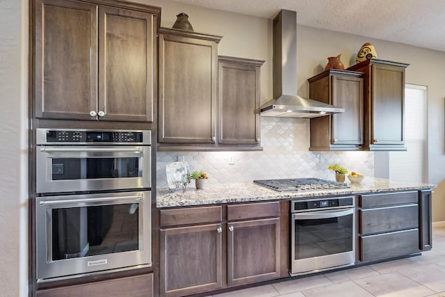 kitchen with stainless steel appliances, wall chimney exhaust hood, light stone counters, and backsplash