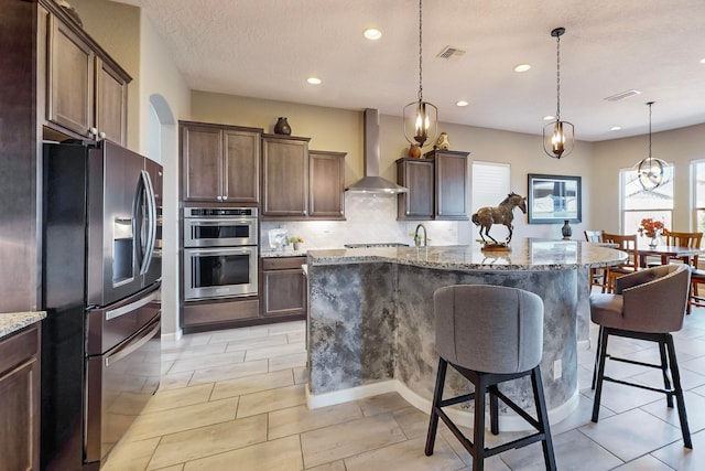 kitchen with an island with sink, light stone countertops, wall chimney exhaust hood, and appliances with stainless steel finishes