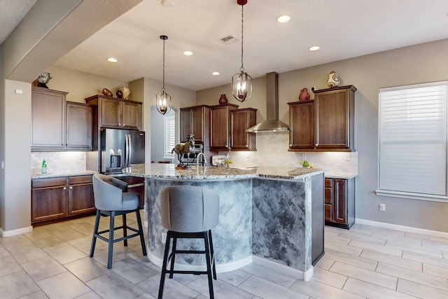 kitchen with light stone counters, an island with sink, stainless steel fridge with ice dispenser, wall chimney range hood, and pendant lighting