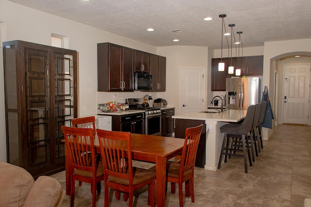 kitchen featuring appliances with stainless steel finishes, a textured ceiling, an island with sink, dark brown cabinets, and pendant lighting