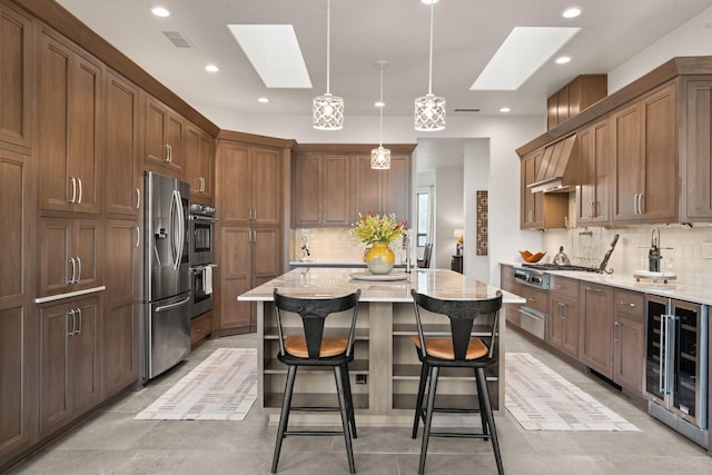 kitchen featuring a skylight, hanging light fixtures, stainless steel appliances, light stone counters, and an island with sink