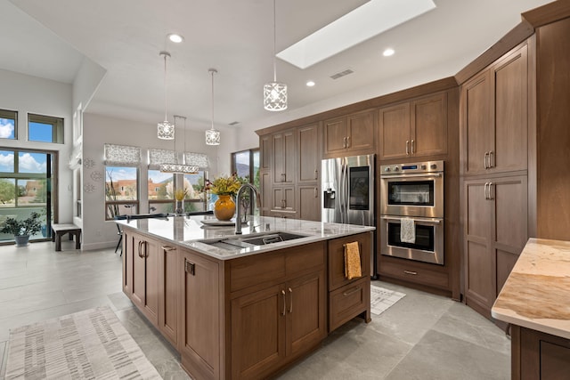 kitchen featuring a kitchen island with sink, sink, hanging light fixtures, light stone countertops, and stainless steel appliances