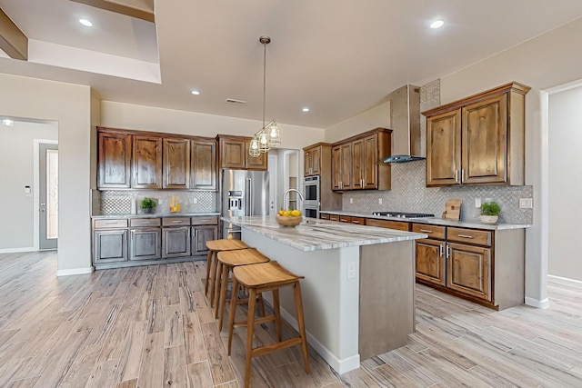 kitchen with stainless steel appliances, an island with sink, light hardwood / wood-style flooring, wall chimney range hood, and decorative backsplash