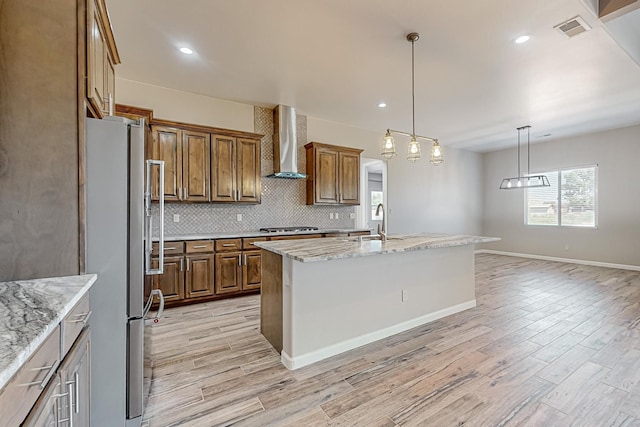 kitchen with light stone counters, stainless steel appliances, light wood-type flooring, an island with sink, and wall chimney exhaust hood