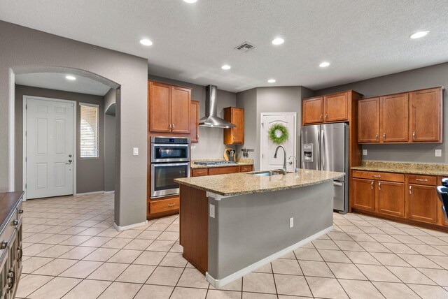 kitchen featuring light tile patterned flooring, wall chimney range hood, appliances with stainless steel finishes, sink, and a kitchen island with sink