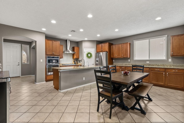 kitchen featuring light stone counters, wall chimney range hood, a kitchen island with sink, and appliances with stainless steel finishes