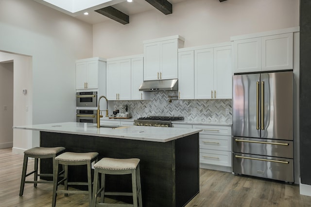 kitchen featuring appliances with stainless steel finishes, a kitchen island with sink, sink, beamed ceiling, and white cabinets