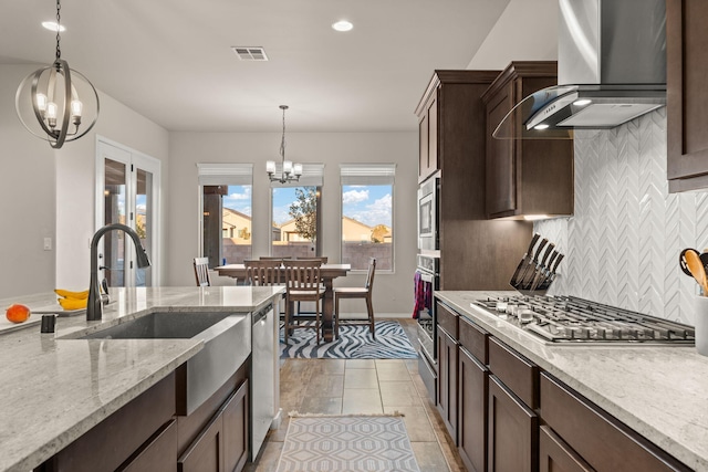 kitchen featuring wall chimney range hood, an inviting chandelier, light stone counters, decorative light fixtures, and appliances with stainless steel finishes