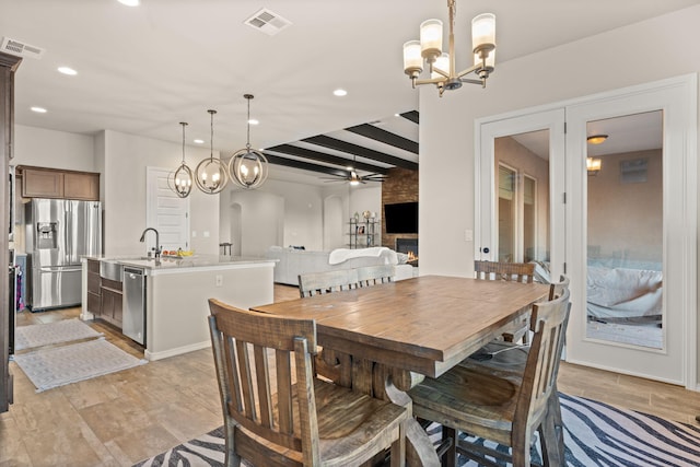 dining space featuring ceiling fan with notable chandelier, sink, light wood-type flooring, beamed ceiling, and a large fireplace